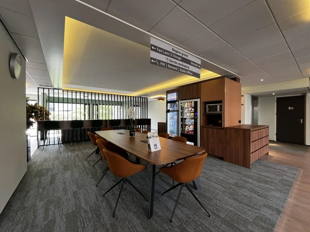 Moderne kantoorlounge met vergadertafel, zitplekken, snackautomaten en bewegwijzering naar onder andere Burgemeester Verderlaan.